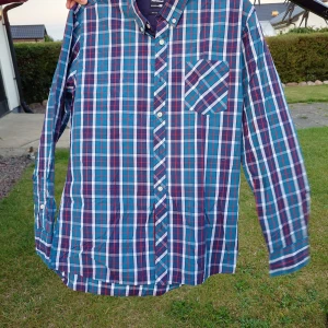 Rutig blå skjorta från Fishbone - Snygg rutig skjorta från Fishbone i blå, vit och röd färg. Slim fit-modell med långärmad design och klassisk krage. Skjortan har knappar framtill och en bröstficka med diagonalt rutmönster. 