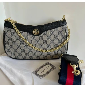 axelväska med kedja och GG-mönster - Snygg axelväska med klassiskt GG-mönster i grått och svart. Väskan har guldfärgad kedja, svarta skinn-detaljer och avtagbar axelrem i blått och rött. Perfekt för dig som vill ha en ikonisk och trendig accessoar.