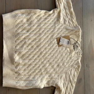 Vit kabelstickad tröja från Aware - Kabelstickad tröja i mjuk vit nyans från Aware. Tröjan har rund halsringning, ribbade muddar och ett klassiskt flätmönster över hela plagget. Perfekt för lager-på-lager och en clean look till jeans eller kjol.