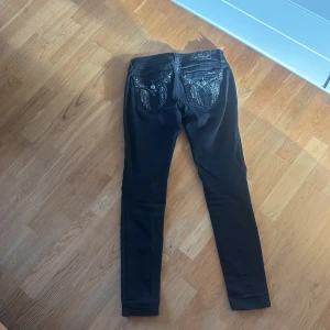 Svarta skinny jeans från Miss Me - Svarta skinny jeans från Miss Me med snygga detaljer som nitar, broderier och glittriga stenar på fickorna. Jeansen har låg midja och smal passform, tillverkade i stretchig denim för en bekväm känsla. Perfekta för dig som gillar att sticka ut med coola detaljer.