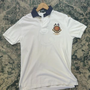 Vit pikétröja Polo Ralph Lauren  - Vit pikétröja från Polo Ralph Lauren i 100% bomull. Klassisk modell med mörkblå krage, broderad logga och emblem på bröstet samt stor siffra 5 på ryggen. Snygg och sportig stil med korta ärmar och knappar vid halsen.