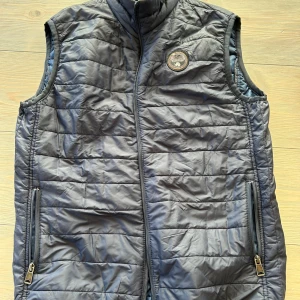 Mörkblå dunväst från Napapijri - Snygg mörkblå dunväst från Napapijri med quiltad design och dragkedja framtill. Västen har två praktiska fickor med dragkedja och en rund logotyp på bröstet. Perfekt för lager-på-lager och stilren look.