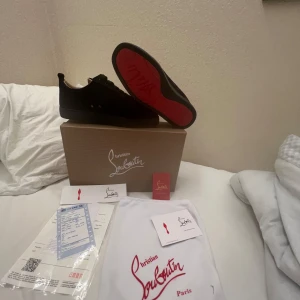 Christian Louboutin svarta sneakers - Exklusiva svarta sneakers från Christian Louboutin med ikonisk röd sula. Skorna har en stilren design i mocka och skinn, rund tå och svarta snören. Kommer med originalkartong, dustbag och äkthetsbevis. Perfekta för dig som vill sticka ut med lyxig streetstyle.
