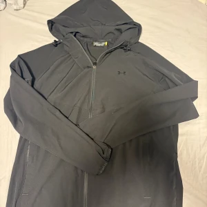 Under Armour vindjacka - Svart vindjacka från Under Armour med huva och justerbar dragsko. Jackan har halv dragkedja framtill, två sidofickor och diskret logga på bröstet. Perfekt för blåsiga dagar och sportiga outfits.