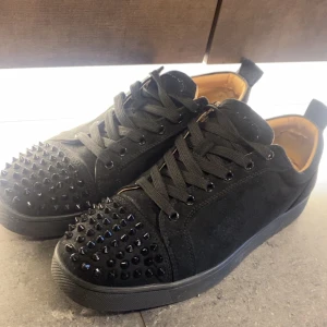 Christian Louboutin  - Svarta sneakers med coola svarta nitar på tån och snörning. Skorna har en platt sula och är tillverkade i mocka med detaljer i syntet. Insidan är fodrad i beige. Perfekta för dig som vill sticka ut med din stil. Ny skick, box följer med 