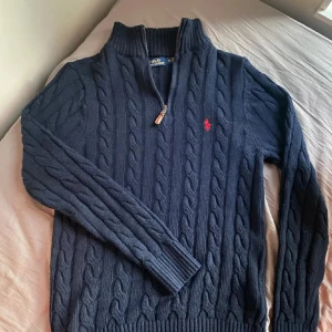 Mörkblå stickad tröja Polo Ralph Lauren - Mörkblå stickad tröja från Polo Ralph Lauren med quarter zip och hög krage. Klassisk röd broderad logga på bröstet. Tröjan är långärmad och sitter ganska slim. Perfekt tröja året runt