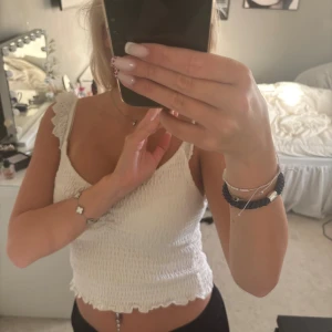 Vit smockad linnetopp från Stradivarius - Supersöt vit linnetopp från Stradivarius med smockad struktur och broderade spetsdetaljer på axelbanden. Toppen har v-ringning och volangkant nertill, perfekt för varma dagar. Lätt och luftig känsla, passar till jeans eller kjol.