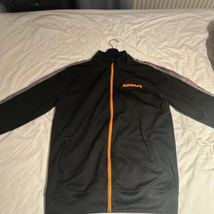 Svart Askari ziptröja med orange detaljer - Svart ziptröja från Askari med orange logga på bröstet och ryggen. Snygga grå ränder med text längs ärmarna och orange dragkedja framtill. Tillverkad i 100% polyester och har två fickor med dragkedja. Perfekt för dig som gillar sportig stil.