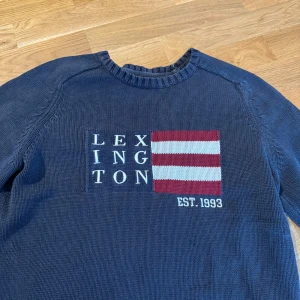 Mörkblå stickad tröja Lexington M - Snygg mörkblå stickad tröja från Lexington med broderad logga och rödvit randig flagga på bröstet. Tröjan har rund halsringning och långa ärmar, perfekt för dig som gillar klassisk och stilren look.
