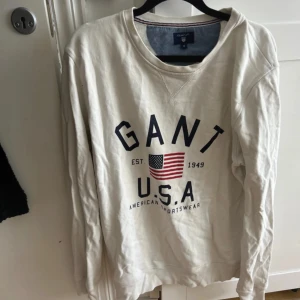 Vit sweatshirt från GANT med tryck - Vit sweatshirt från GANT med stort blått och rött USA-tryck framtill och amerikansk flagga. Klassisk rund halsringning med ribbade muddar och insida i mjuk bomull. Perfekt för en avslappnad och sportig stil.