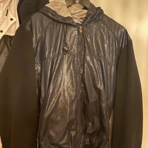 Svart vindjacka från EA7 Emporio Armani - Snygg svart vindjacka från EA7 Emporio Armani med glansig framsida, huva med snörning och dragkedja. Jackan har stora EA7-loggan på ryggen och mönstrat innerfoder med logotyper. Perfekt för dig som vill ha en sportig och stilren look. Passar M