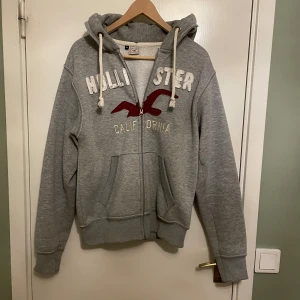 Grå hoodie från Hollister med dragkedja - Grå hoodie från Hollister med broderad logga och text på bröstet. Tröjan har huva med snörning, dragkedja framtill och två fickor. Mjuk insida och klassisk passform, perfekt för chill dagar.  Skick som på bilderna och vid frågor skriv privat!