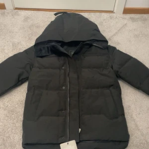 Canada Goose jacka - Säljer en svart dunjacka från Canada Goose med huva och dragkedja. Jackan är vadderad och har en klassisk, stilren look med diskreta detaljer. Perfekt för kalla dagar och riktigt varm. Passar dig som vill ha en snygg och funktionell vinterjacka.