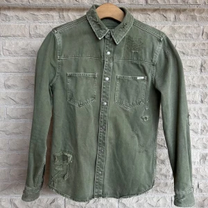 Grön jeansskjorta med slitna detaljer - Säljer en grön jeansskjorta med coola slitna detaljer. Skjortan har långa ärmar och två bröstfickor med knappar. Perfekt för en avslappnad och trendig look. Passar bra till både vardag och fest!