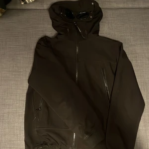 Svart Goggle Jacket från C.P. Company - Svart Goggle Jacket från C.P. Company med ikoniska glasögon i huvan. Jackan har dragkedja framtill, flera fickor med dragkedja och är tillverkad i ett slitstarkt syntetmaterial. Perfekt för dig som vill sticka ut med en streetwear-inspirerad look. Strlk M men passar även folk med strlk S! Låga priset pågrund av ganska risigt skick. Ena glaset är sprucket och en del styng är lösa. 