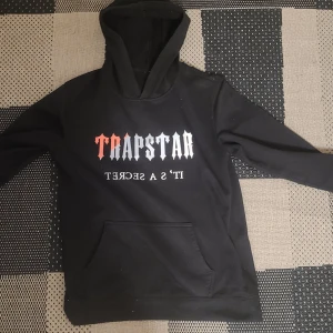 Svart Trapstar hoodie med tryck - Svart hoodie från Trapstar med stor huva och magficka. Framsidan har ett iögonfallande tryck i vitt och rött med texten 'TRAPSTAR' och 'IT'S A SECRET'. Perfekt för dig som gillar streetwear och vill sticka ut.