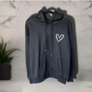 Svart hoodie med hjärta från SHEIN - Svart hoodie från SHEIN i storlek S, tillverkad i 100% polyester. Tröjan har en stor huva med snörning, magficka och ett vitt hjärta tryckt på bröstet. 