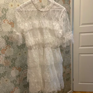 Vit spetsklänning från Bubbleroom - Superfin vit kortklänning från Bubbleroom i spets med volangdetaljer och hög hals. Klänningen har korta ärmar och knäppning i nacken. Endast använd 1 gång, men har lite sminkfläckar vid halsringningen och i nacken. Tror absolut det går bort med sminkborttagning eller i tvätten 