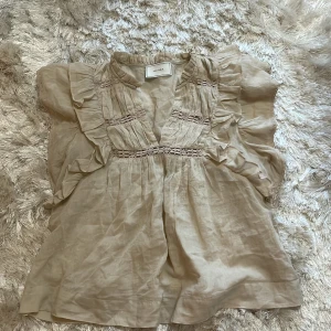 Beige blus med volang från Neo Noir - Supersöt beige blus från Neo Noir med volangdetaljer längs axlar och bröst. Blusen har v-ringning och fina spetsinslag framtill. Luftigt material och romantisk känsla, perfekt för vår och sommar. Passar till både jeans och kjol.