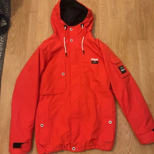 Röd vindjacka från Dope med huva - Snygg röd vindjacka från Dope med stor huva och vita snören. Jackan har flera praktiska fickor, svart innerfoder och coola logodetaljer på bröstet och ärmen. Perfekt för dig som vill sticka ut med en färgstark och funktionell jacka.