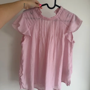Ljusrosa blus med volangärm - Supersöt ljusrosa blus med volangärmar och rynkade detaljer framtill. Blusen har rund halsringning och är tillverkad i ett lätt, luftigt tyg som känns skönt mot huden. Perfekt att styla med jeans eller kjol för en fräsch look.