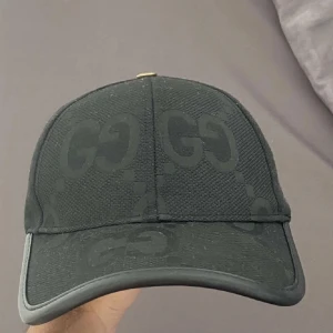 Svart  keps med GG-mönster - Snygg svart keps med diskret GG-logomönster över hela kepsen. Kepsen har böjd skärm och justerbar rem bak med metallspänne. Materialet är en mix av bomull och polyester, vilket gör den både bekväm och stilren. Perfekt för dig som vill ha en lyxig streetwear-look.