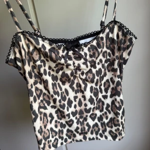 Leopardmönstrad linne från Gina Tricot - Snygg leopardmönstrad linne från Gina Tricot med tunna axelband och svart spetskant upptill. Toppen har en liten rosett framtill och är i mjukt, stretchigt material. Perfekt för dig som vill sticka ut med ett djurmönster. Använd en gång.