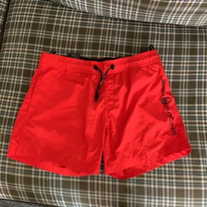 Röda Sail Racing badshorts med snörning - Riktigt snygga röda badshorts från Sail Racing med svart logga på ena benet. Shortsen har elastisk midja med svart snörning och en bakficka med Sail Racing-märke. Perfekta för stranden eller poolen, tillverkade i snabbtorkande syntetmaterial.