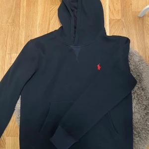 Svart hoodie från Polo Ralph Lauren - Svart hoodie från Polo Ralph Lauren med klassisk röd broderad logga på bröstet. Tröjan har huva, magficka och långa ärmar. Tillverkad i mjuk bomull och perfekt för chill dagar. Enkel och stilren design som funkar året runt.