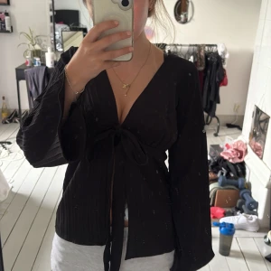 Svart omlottströja  - Säljer en svart omlottopp med långa ärmar, Toppen har knytband framtill och är tillverkad i ett lätt, plisserat tyg som ger en snygg struktur. Köpte tröjan i Italien för cirka 1 år sen och har aldrig använt den. Passar Xs/S