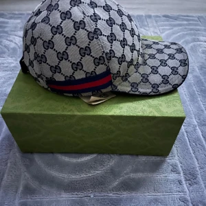 Gucci keps med GG-mönster, storlek L - Exklusiv keps från Gucci i marinblåa och svart med klassiskt GG-monogram över hela kepsen. Kepsen har en böjd skärm och en ikonisk röd-blå rand på sidan. Tillverkad i bomullsmix och har justerbar rem baktill för perfekt passform.