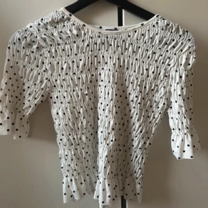 Prickig smockad blus från Gina Tricot - Supersöt vit blus från Gina Tricot med svarta prickar och smockad design. Blusen har korta ärmar med volangkant och en figurnära passform. Perfekt för dig som gillar trendiga och lekfulla detaljer.