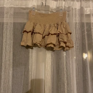 Beige volangkjol från SHEIN - Supersöt beige kortkjol från SHEIN med flera lager volanger och smockad midja. Kjolen är lätt och luftig, perfekt för varma dagar. Enkel att matcha med crop tops eller stickade tröjor för en trendig look. Endast prövad