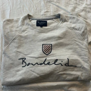Ljusgrå sweatshirt från Bondelid - Snygg ljusgrå sweatshirt från Bondelid med stort tryck på bröstet och ett randigt emblem. Klassisk rund hals och mjukt bomullsmaterial som är skönt mot huden. Perfekt för dig som gillar stilrena och enkla tröjor med en modern touch.