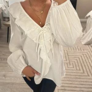 Vit blus med volang och snörning - Supersöt vit blus med v-ringning, volangdetaljer längs halsringningen och snörning framtill. Blusen har långa, luftiga ärmar med resår vid handlederna och är tillverkad i ett lätt bomullstyg. Perfekt för dig som gillar romantisk och trendig stil.