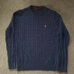 Blå kabelstickad tröja från Morris - Snygg kabelstickad tröja från Morris i marinblått. Tröjan är gjord av 100% pima bomull och har ett broderat emblem på bröstet. Rund halsringning och långa ärmar med ribbade muddar. Perfekt för lager-på-lager under kyliga dagar. Bra skick. Nypris cirka 1700-1800