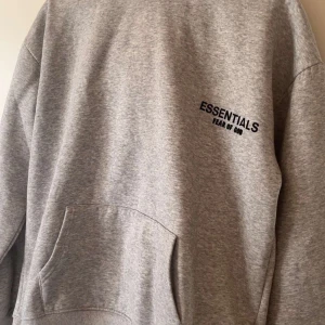 Grå Essentials Fear of God hoodie - Säljer en grå hoodie från Essentials Fear of God med svart logga på bröstet och ryggen. Tröjan har huva, känguruficka och är i mjuk bomullsblandning. Perfekt för dig som gillar streetwear och oversized look.