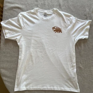 Vit Quiksilver t-shirt California - Helt o använd vit t-shirt från Quiksilver med stort tryck på ryggen där det står 'Quiksilver California' och en björn i brunt och vitt. Liten björnlogga på bröstet. Tillverkad i 100% bomull, perfekt för en chill och avslappnad stil. Bara hör av er om ni har några frågor!