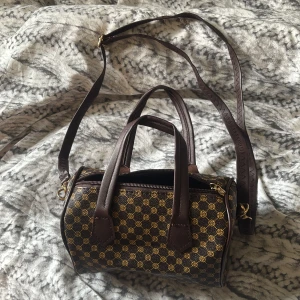 Brun axelväska med mönster - Snygg brun axelväska med klassiskt mönster i gult och brunt. Väskan har både handtag och justerbar axelrem samt dragkedja upptill. Perfekt storlek för vardagens essentials och ger en lyxig känsla med sina guldfärgade detaljer.