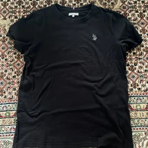 Svart t-shirt från U.S. Polo Assn - Svart t-shirt från U.S. Polo Assn. med klassisk passform och rund hals. T-shirten har korta ärmar och en broderad logga på bröstet. Tillverkad i mjuk bomull som är skön att bära hela dagen.