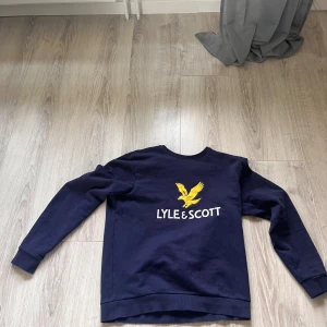 Mörkblå Lyle & Scott sweatshirt - Säljer en mörkblå sweatshirt från Lyle & Scott med stor gul broderad örn och vit logga på bröstet. Tröjan har rund hals, långa ärmar och ribbade muddar. Perfekt för dig som gillar klassisk streetstyle och vill ha något bekvämt och snyggt.