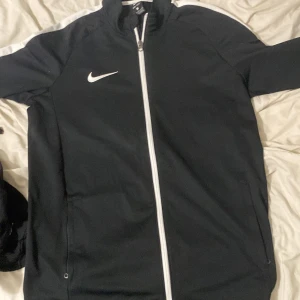 Nike svart kofta Dri-Fit - Svart Nike kofta med vit dragkedja och vit logga på bröstet. Koftan har vita detaljer längs axlarna, sidofickor med dragkedja och är tillverkad i Dri-Fit polyester som andas. Perfekt för träning och chill. Långärmad och normal passform.