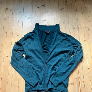 Mörkgrön kofta från Oscar Jacobson - Mörkgrön kofta från Oscar Jacobson med dragkedja framtill och hög krage. Tillverkad i Italien och i ett tunt, stickat material som känns mjukt mot huden. Perfekt för lager-på-lager och snygg till hösten.