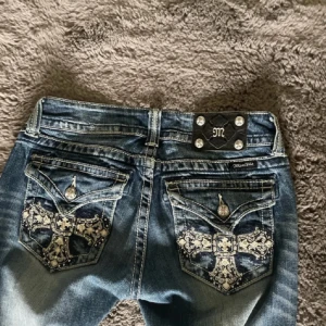 Miss Me straight jeans med broderi - Snygga blå Miss Me jeans i straight fit med slitningar och broderade bakfickor med korsdetaljer och bling. Jeansen har silverfärgade knappar och detaljer, samt coola slitningar framtill. Perfekt för dig som gillar statement-jeans med extra detaljer.