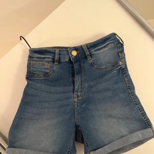 Blå jeansshorts från Gina Tricot - Snygga blå jeansshorts från Gina Tricot med klassisk femficksdesign och uppvikta benslut. Modellen är högmidjad och har en knapp och dragkedja framtill. Perfekt för varma dagar och enkel att matcha med din favorit-topp.