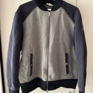 Grå och marinblå bomberjacka Moncler - ÄKTA Snygg bomberjacka från Moncler i grått och marinblått med klassisk logga på ärmen. Jackan har ribbade muddar, två fickor med knappdetalj och dragkedja framtill med dubbla zippar. Perfekt för dig som vill ha en stilren och sportig look. Tröjan är true to size, men passar även dig med storleken L
