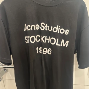 Svart Acne Studios t-shirt Stockholm - Svart t-shirt från Acne Studios med vit text på ryggen: 'Acne Studios Stockholm 1996'. Klassisk passform och rund hals. Perfekt basic med cool print för dig som gillar streetstyle och skandinavisk design.