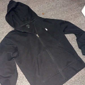 Svart hoodie från Polo Ralph Lauren - Svart hoodie från Polo Ralph Lauren med dragkedja framtill och klassisk vit logga på bröstet. Tröjan har huva, två fickor och långa ärmar. Perfekt för en chill och stilren look.