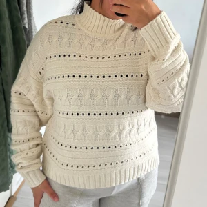 Vit stickad polotröja med kabelmönster - Mysig vit stickad polotröja med bred ribbad krage och ärmslut. Tröjan har ett klassiskt kabelstickat mönster och dekorativa hål för en extra touch. Perfekt för kyliga dagar och enkel att matcha med jeans eller mjukisbyxor.