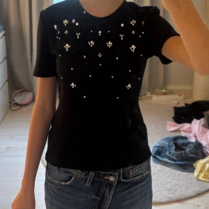 Svart topp med pärldetaljer från Zara - Säljer en svart topp med korta ärmar och rund halsringning. Toppen är dekorerad med vita pärlor i blommönster framtill, vilket ger en snygg och unik look. Perfekt att styla med jeans eller kjol för en trendig outfit.